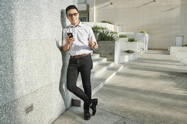 オフィスビルに立ち 親指を立ててスマートフォンを持ったサングラスをかけた陽気なベトナム人ビジネスマン — ストック写真