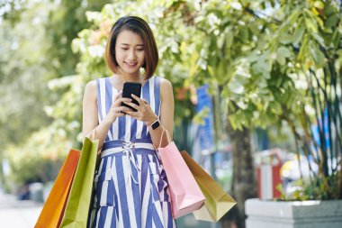 Alışveriş poşetlerini tutan Asyalı genç kadın cep telefonunda bir mesaj okuyarak ve sokakta yürürken gülümseyerek