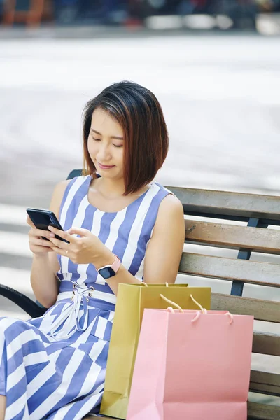 短い髪のアジアの若い女性は 彼女の近くに紙の買い物袋を持ってベンチに座って 彼女は買い物の後に休んでいる彼女の携帯電話を使用しています — ストック写真