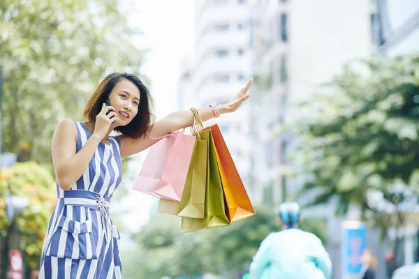 ショッピングバッグを着た美しいアジアの女性は 市内の屋外でタクシーをキャッチしながら 携帯電話で会話を持っています — ストック写真