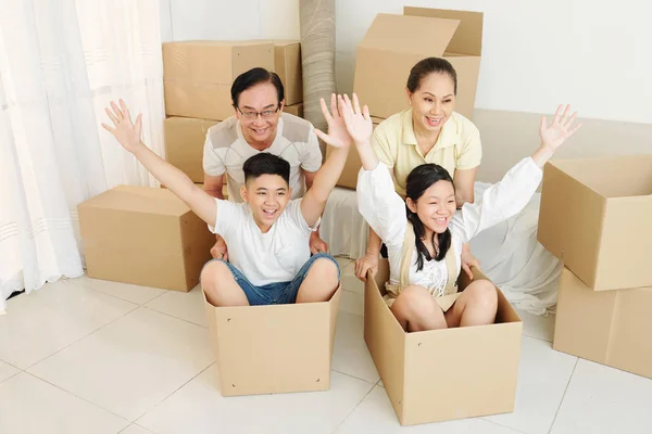 Happy grandparents pushing cardboard boxes with grandchildren sitting inside after moving to new house