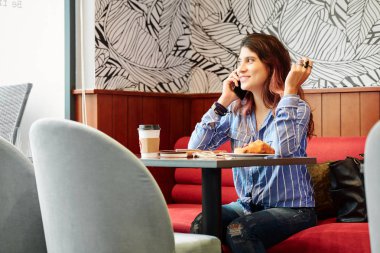 Kafe masasında oturan, pencereden bakan ve telefonla konuşan pozitif, güzel bir kadın.