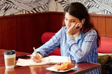 Kafe masasında oturan, kulaklıkla ders dinleyen ve önemli düşünceler yazan genç ve sıkkın bir kadın.