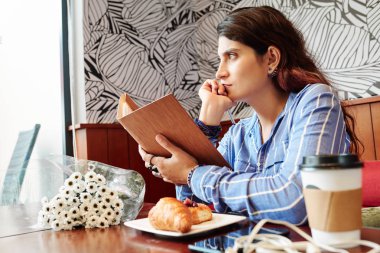 Elleri açık kafeterya masasında çiçek buketi, hamur işleri, kahve ve pencereden bakan düşünceli genç bir kadın.