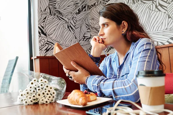 一个多愁善感的年轻女人 手里拿着翻开的书 坐在茶几旁 手里拿着花束 糕点和咖啡 望着窗外 — 图库照片