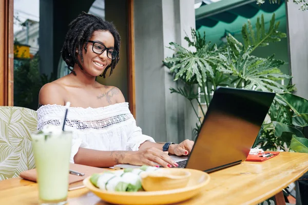 戴着眼镜 面带微笑的非洲女商人坐在桌旁 一边吃着东西 一边在户外咖啡店的笔记本电脑上打字 — 图库照片