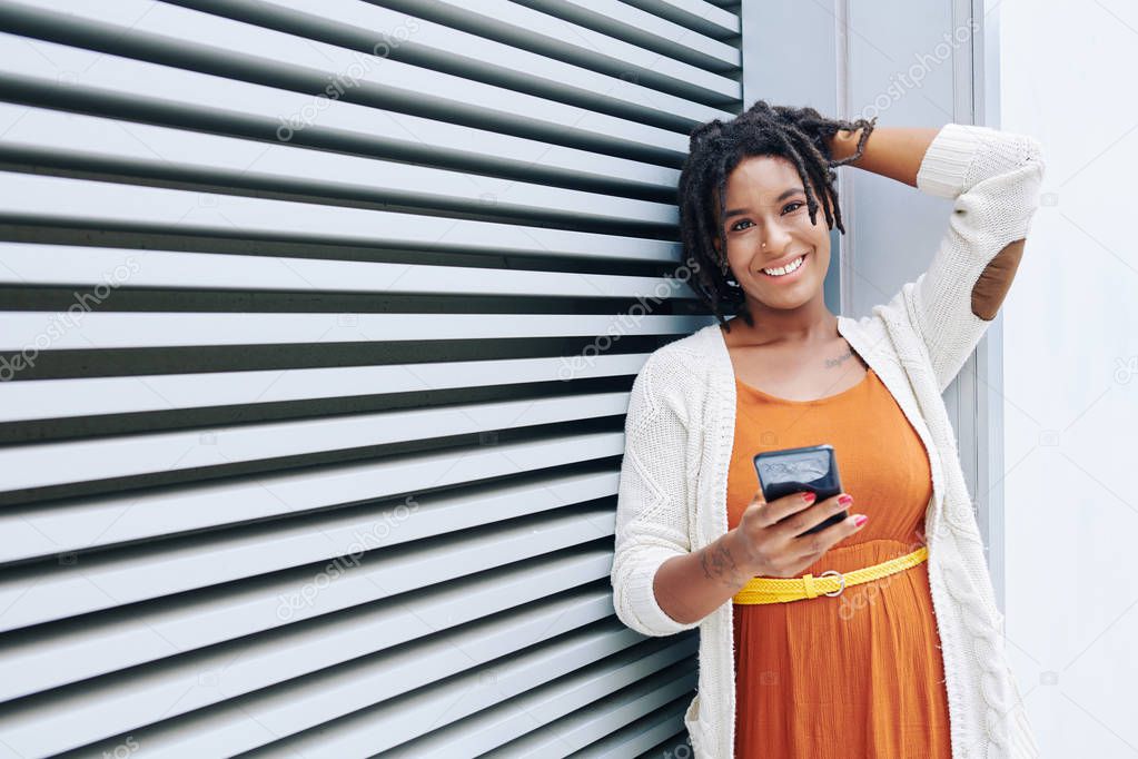 Portrait of African young woman in orange dress standing with mobile phone and smiling at camera against the striped wall outdoors