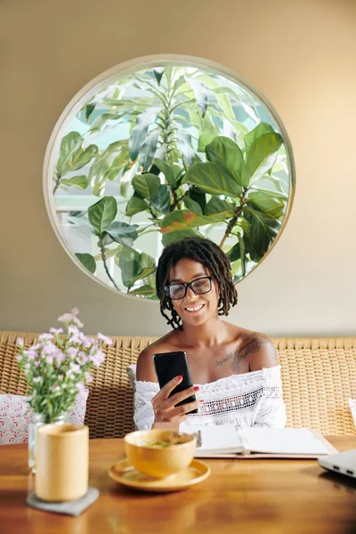 戴着眼镜的美丽的非洲女人一边在咖啡店吃午饭 一边在手机上看信息 微笑着 — 图库照片