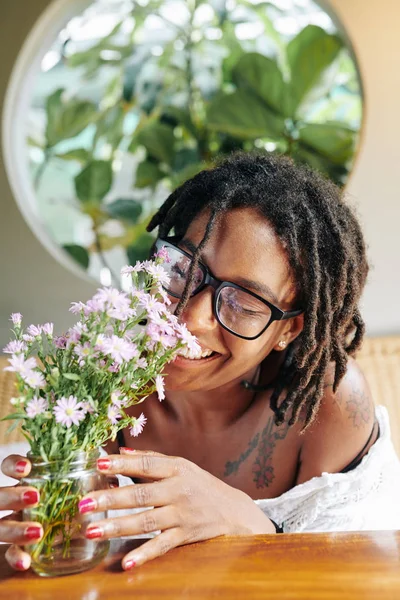 Happy African woman in eyeglasses and with beautiful hairstyle smelling flowers in the vase on the table and smiling