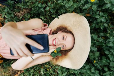 Parktaki yeşil çimlerin üzerinde yatarken fotoğraf çeken güzel, güler yüzlü bir kızın görüntüsünün hemen üstünde.