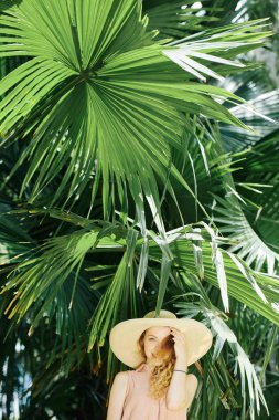 Hasır şapkalı, genç ve güzel bir kadın yaz günü palmiye ağacının altında duruyor.
