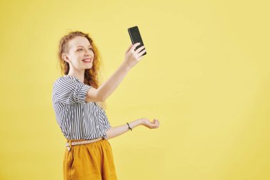 Neşeli, güzel, günlük kıyafetler içinde, gülümseyen ve akıllı telefondan fotoğraf çeken genç bir kadın.