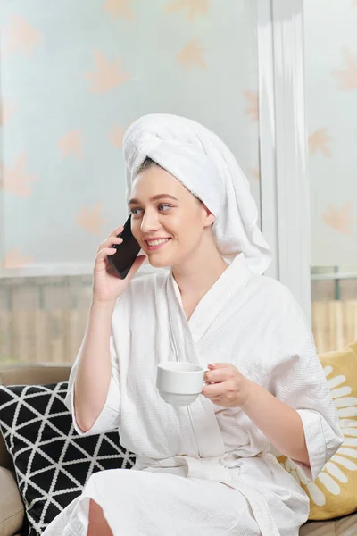 Pretty young woman in bath robe and towel on her head drinking morning coffee and chatting on phone