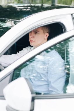 Genç, ciddi bir adam beyaz arabasının direksiyonunda otururken önüne bakıyor.