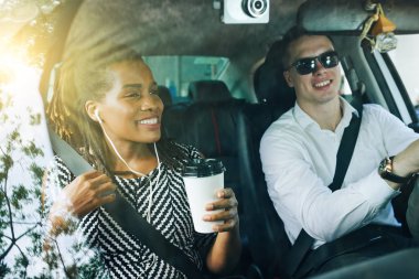 Afrikalı genç bir kadın kahve içerken gülümsüyor ve araba sürerken genç bir adamla konuşuyor.
