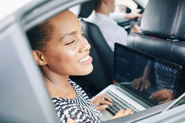 African smiling woman sitting on back seat and using laptop computer while driving
