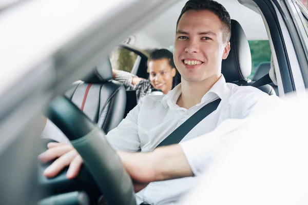 後部座席で女性乗客と車を運転しながらカメラで微笑む若い男の肖像画 — ストック写真