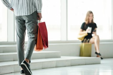 Alışveriş poşetleriyle alışveriş merkezinde oturan ve alışveriş yaptıktan sonra arkadaşlarına mesaj atan genç bir adam.