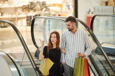 Güzel genç bir kadın kocasıyla yürüyen merdivende dururken alışveriş merkezinde butiği işaret ediyor.