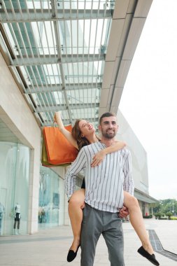 Neşeli yakışıklı genç adam alışveriş merkezinden birlikte alışveriş yaptıktan sonra kız arkadaşını sırtında taşıyor.