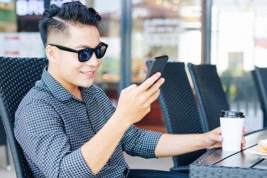 Güneş gözlüklü gülümseyen adam kafede kahve içiyor ve akıllı telefonuyla sosyal medyayı kontrol ediyor.