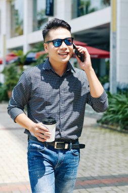 Pozitif genç Asyalı adam dışarıda yürüyor, telefonla konuşuyor ve elinde kahve tutuyor.