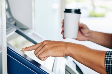 Elinde bir fincan kahveyle bankaha makinesinin şifresini girerken ve banka hesabından para çekerken yakın plan görüntüsü.