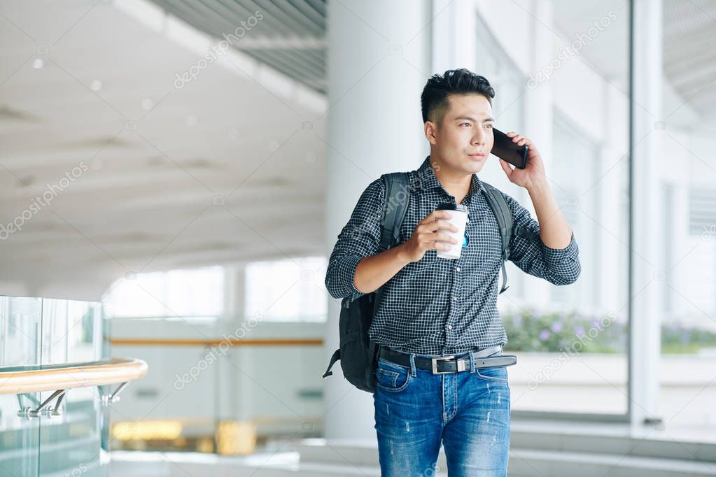 Handsome young Vietnamese man with backpack carrying cup of take away coffee and talking on phone when standing in office building