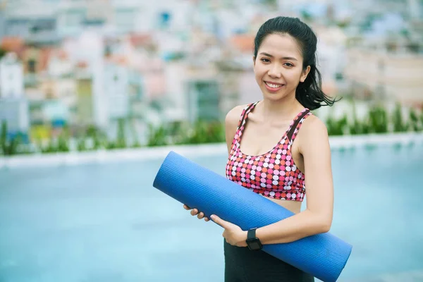屋外プールサイド近くに立ちながら運動マットを持ちカメラで微笑むアジアの若い女性の肖像 — ストック写真
