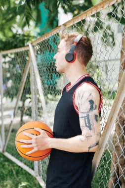 Yakışıklı genç adam sokak basketbol sahasında ağlara yaslanıyor ve çeyreklikler arasında dinlenirken kulaklıkla müzik dinliyor. 