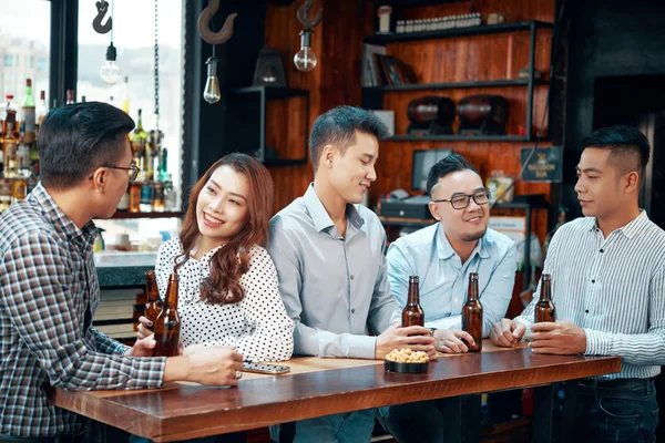 Group of Asian young people talking to each other drinking beer from the bottles while standing at bar counter in the pub