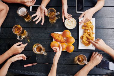Ahşap masada oturan ve bira içip fast food yerken el kol hareketi yapan gençlerin yüksek açılı görüntüsü