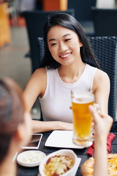 亚洲的年轻女子在餐桌边对着她的朋友笑 而她们则在餐馆边喝啤酒边互相交谈 — 图库照片