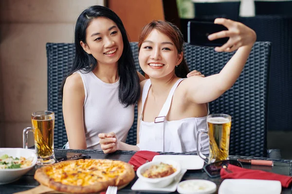 当他们在咖啡店里吃披萨和喝啤酒时 快乐的亚洲女人和她的朋友一起在手机上自画像 — 图库照片