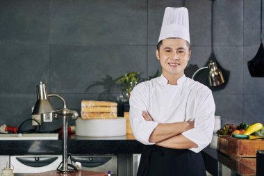 Asyalı genç aşçının portresi. Kollarını kavuşturup kameraya gülümsüyor.