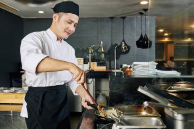 Asyalı genç aşçı sobanın yanında duruyor ve restoranın mutfağında özel yemek için pilav pişiriyor.
