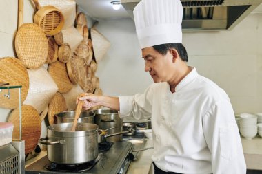 Üniformalı Asyalı olgun şef sobanın yanında duruyor ve mutfaktaki tavada çorbayı karıştırıyor.