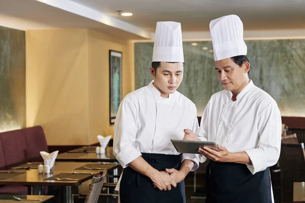 两名厨师站在餐厅时 身穿制服 头戴厨师帽 一起使用数码平板电脑 — 图库照片