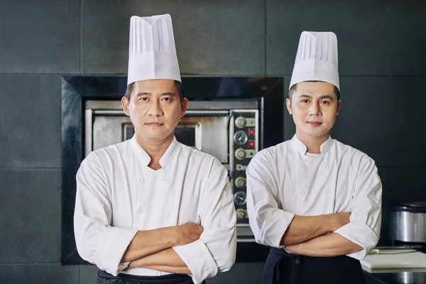 亚洲成熟厨师和他的助手手挽手站在一起 看着厨房里的相机的画像 — 图库照片