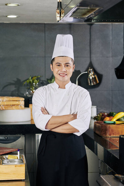 Portrait of Asian young chef in uniform smiling at camera while standing in commercial kitchen of the restaurant