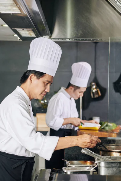 Asyalı Olgun Aşçı Sobanın Yanında Duruyor Mutfakta Asistanıyla Birlikte Yemek — Stok fotoğraf