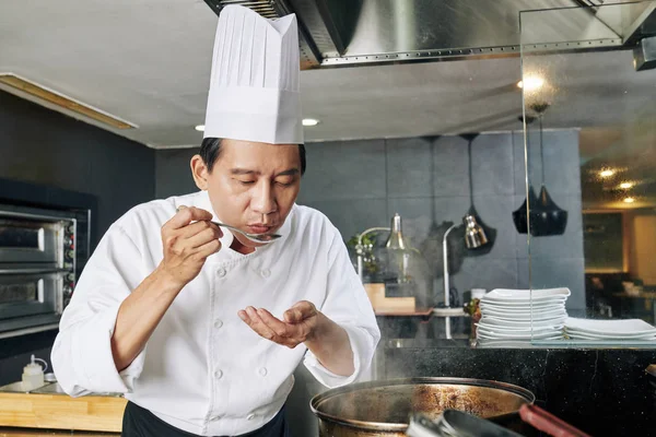 亚洲成熟的厨师头戴白帽 站在炉边 用平底锅品尝刚在厨房准备的汤 — 图库照片