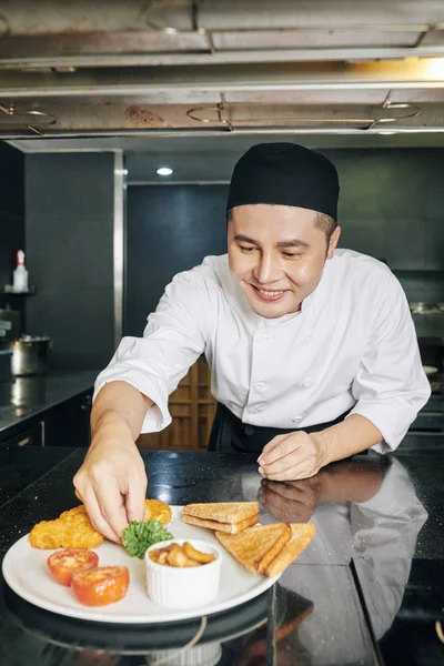 亚洲的年轻厨师在餐馆的厨房里用绿色装饰着他准备好的菜 面带微笑 — 图库照片