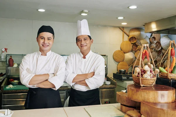 两名身穿制服的亚洲厨师在商业厨房工作时 双臂交叉 对着相机笑着的画像 — 图库照片