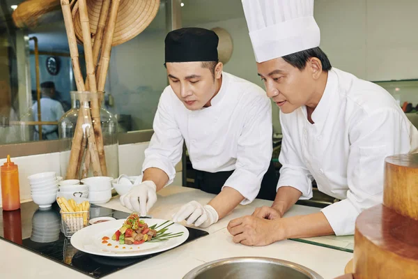亚洲成熟的厨师教他的学生在餐馆的桌子上端菜 — 图库照片