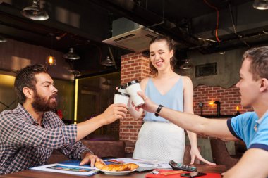 Bir grup güler yüzlü yaratıcı yönetici kafeterya masasının üzerinde doküman ve atıştırmalıklarla kahve içip kadeh kaldırıyor.