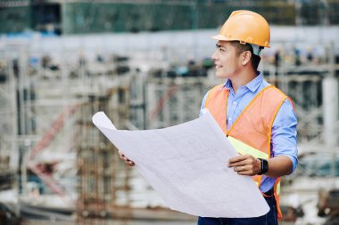 Elinde bina planıyla gülümseyen Asyalı müteahhit inşaat sahasına bakıyor ve çalışma dizisini inceliyor.