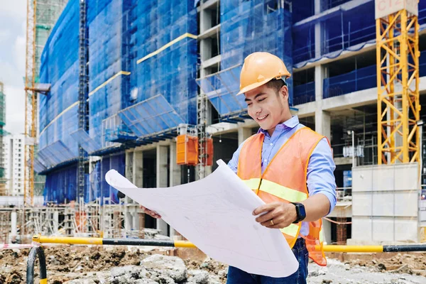 オレンジベストでアジアの建設技術者を笑顔と建物の青写真を検討するハード帽子 — ストック写真
