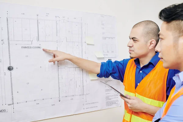 Serious engineer with digital tablet in hands pointing at building blueprint hanging on wall and discussing work