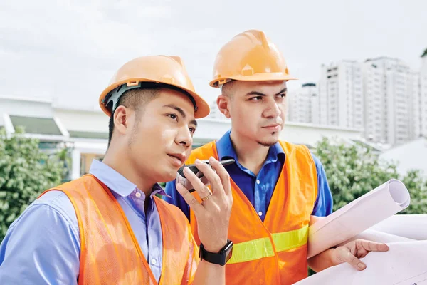越南承包商与工人谈论对讲机 他的同事手握蓝图站在旁边 — 图库照片